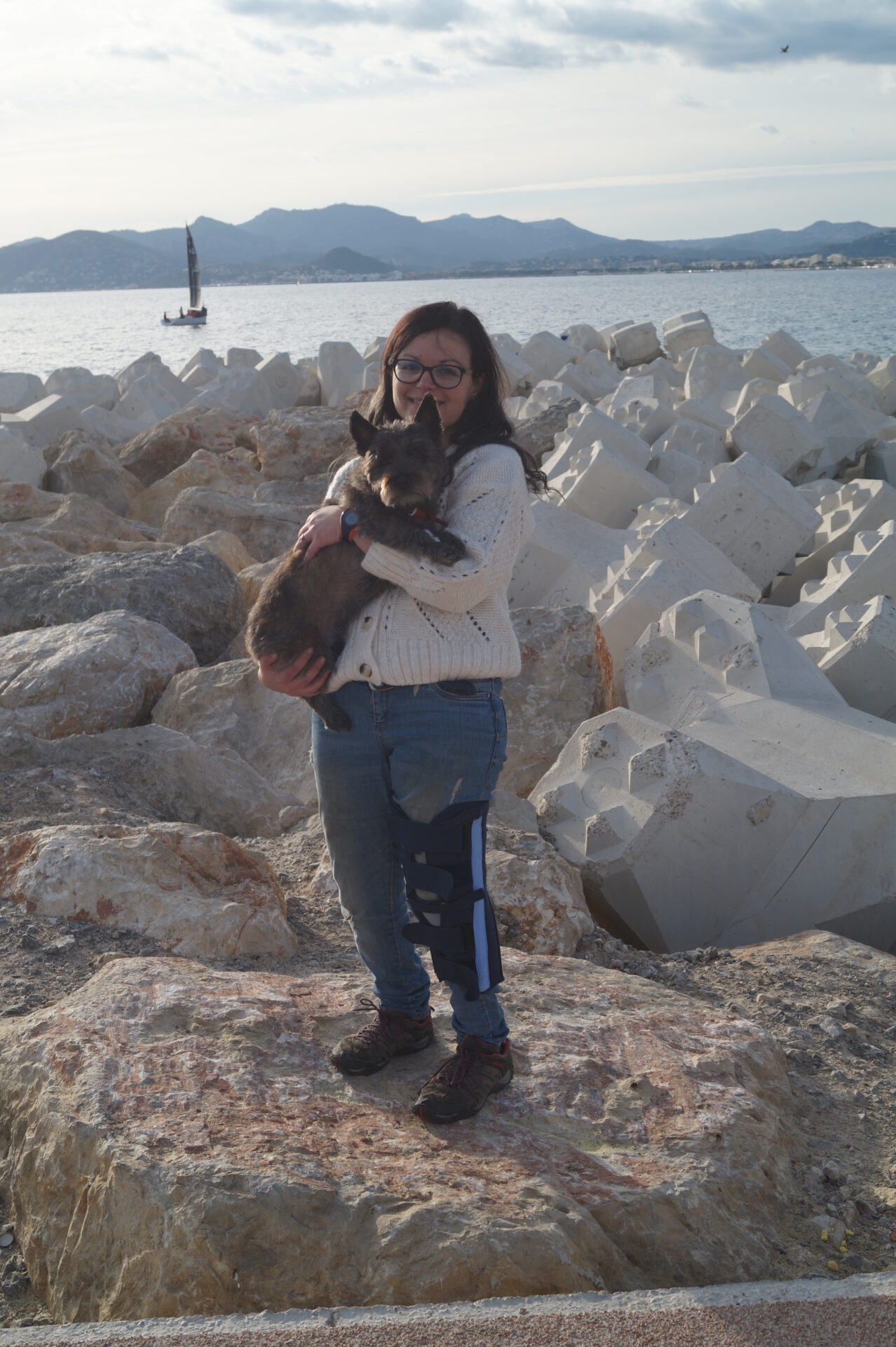 Une photo de Lauriane avec une atèle à la jambe et sa chienne dans ses bras sur le bord de mer de Cannes